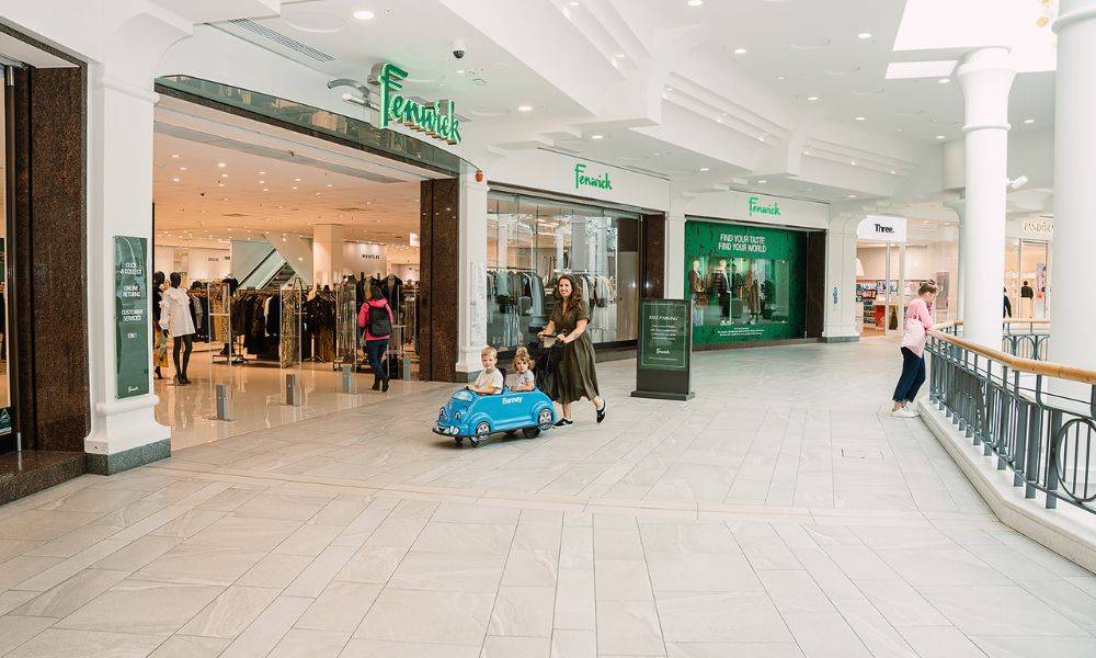 Woman pushing children in a small push-along car next to department store in a shopping centre.