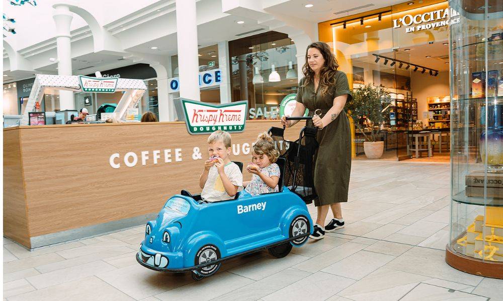 Mother walking with children, eating doughnuts, in a small push-along car in a shopping centre
