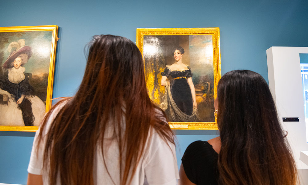 The-Camden-Family-at-the-Amelia two ladies looking the portraits in the Camden Room at the Amelias Scott. The portrait are placed on a Royal Blue wall