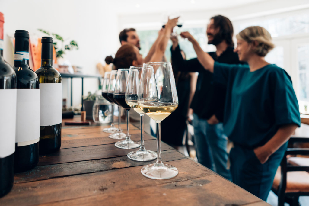 Wine-tasting group of people tasting wine. The foreground includes a line of wine glasses and various bottles with labels covered.
