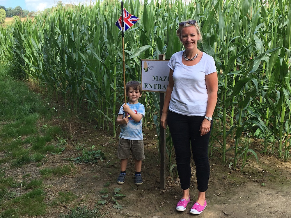 A woman and her child stood smiling at the beginning of a Maize Maze entrance with tall green Maize stood behind them