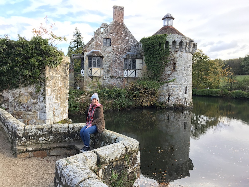 A woman smiling, wearing a hat and scarf sat on a wall in front of Scotney Castle and its moat in the background